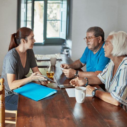A young woman sitting at a wooden table across from an older couple, holding a tablet and gesturing during a conversation.