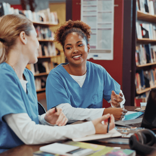Two students in blue medical scrubs studying together at a library table with books and a laptop.