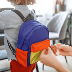 A close-up of a child wearing a colorful blue, orange, and yellow backpack while an adult's hands attach a white tracking tag on a carabiner to it.