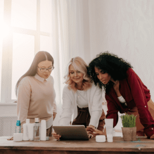 Three female colleagues standing at a wooden desk lined with bottles while reviewing content together on a tablet.