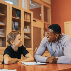 A smiling educator leaning over a table to talk with a young female student in a library or classroom.