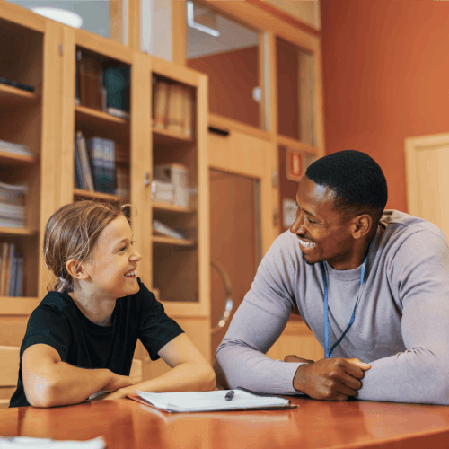 A smiling educator leaning over a table to talk with a young female student in a library or classroom.